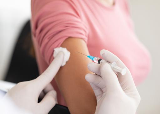 Médecin ayant des gants blancs, qui vaccine à l'aide d'une seringue le bras d'une patiente possédant un haut rose